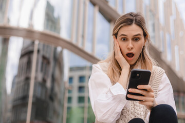 Sad being victim of cyber bullying online, anxious women checks the phone, reads a message on the stairs outside from the business center. Woman shocked and clutched her head, open mouth.