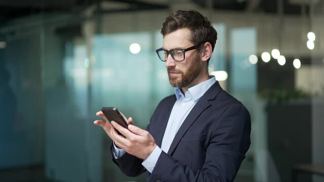 Handsome businessman in formal suit uses mobile phone in modern glass office Bearded male in glasses typing browsing chatting smartphone at workplace Indoor confident entrepreneur financier or broker