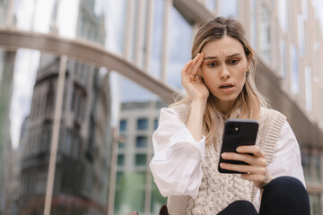 Sad being victim of cyber bullying online, anxious women checks the phone, reads a message on the stairs outside from the business center. Woman shocked and clutched her head.