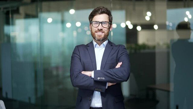 Portrait of successful mature boss, senior businessman bearded man in glasses and business formal suit looking at camera and smiling, man with crossed arms working inside modern office building indoor