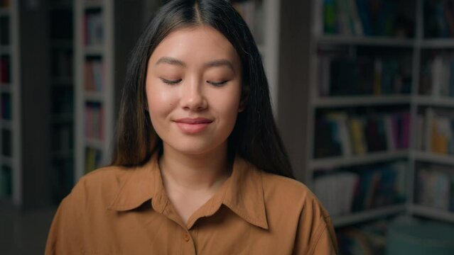 Close-up happy asian young woman posing in library looking at camera girl student sitting at desk with books read preparing for university exam studying homework book reading hobby moving footage blur