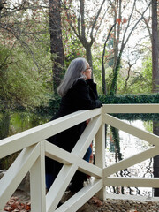 Fototapeta premium Older woman with gray hair and glasses next to a tree feeling it. Woman leaning on the trunk of a tree in a park with green grass. Girl in prairie and green forest pensive and nostalgic