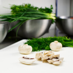Fresh sliced mushrooms on a cutting board. In the background are green onions, parsley and dill in a stainless steel bowl