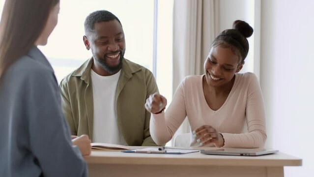 Young african american man and woman signing contract, real estate agent giving them keys from new apartment