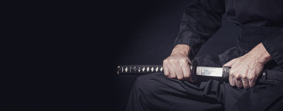 The samurai holding a Japanese katana sword, banner. Photo of a warrior dressed in black clothes in low key with copy space for text