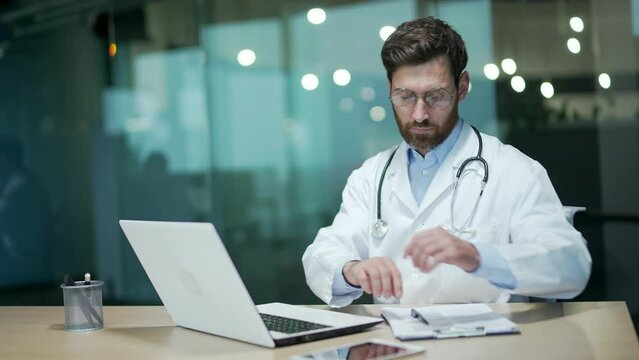 Confident busy mature doctor therapist with stethoscope looking at laptop computer screen typing browsing working online distance remote and make notes in patient's medical card at modern hospital