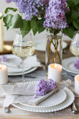 Beautiful table decor for a wedding dinner with a spring blooming lilac flowers. Celebration of a special holiday marriage event. Fancy white plates, wineglasses, candles. Countryside style