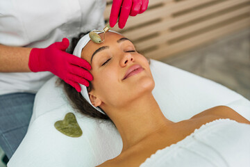 Naklejka premium Hands of the beautician massaging clients face with a jade roller, woman lying in a spa salon on a massage table
