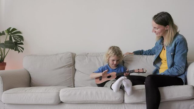 Happy kid son playing guitar ukulele with mother at home living room - Family, music concept