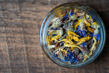 Dry flower and herbal tea leaves in a glass jar on wooden background. Herbal collection of chamomile, cornflower, mint, sea buckthorn, lemongrass, wild rose, dried citrus fruits and apple