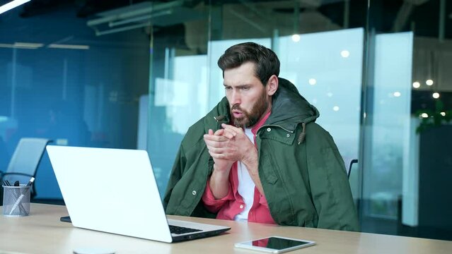 Cold bearded business man freelancer covered by jacket suffers and trembles warms hands try to work on laptop computer at desk in modern office with problem of heating system indoors Low temperature
