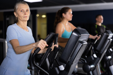 Fototapeta na wymiar Portrait of motivated sporty senior woman doing cardio training, exercising on elliptical trainer in gym
