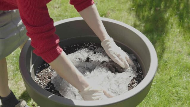 mixing soil with hydro-gel nanotechnology granules for planting flowers in big pot outdoors sunny day. red female hands in garden gloves add colloidal consistency polymer reducing irrigation into