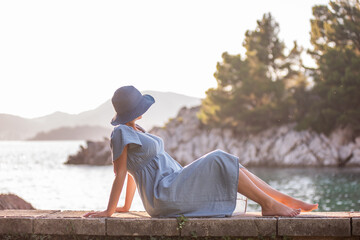 Tender pregnant woman in denim dress, blue hat sits by the sea in the sun, backdrop of green rock island. Traveling in pregnancy to Montenegro on the Royal Beach. Mothers care rest vacation. Botanical