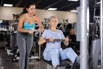 Young female trainer giving advice to old lady training at cable row machine
