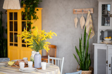 Spring, Easter interior in Scandinavian style. Rustic Living room with a bright wardrobe, yellow mimosa in vase. The table is festively set with ceramic dishes, with a cake on a tray. macrame details