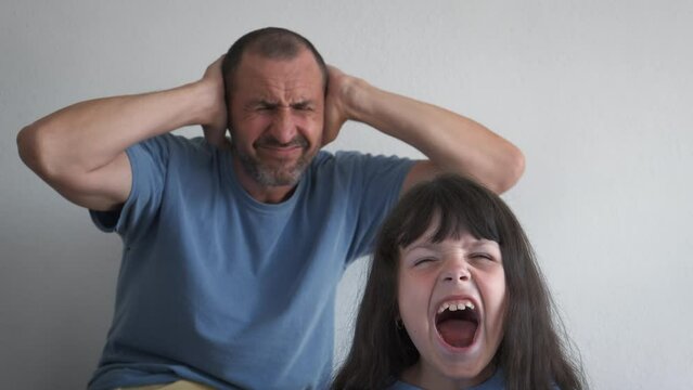 A naughty child screams loudly. The hysterical little daughter screams, the father covers his ears with his hands from stress.