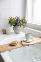 Cup of coffee, candles, book and soap on wooden board. Elegant Scandinavian bathroom interior with white tiles.Blurred background. Colorful tulips, hyacinth spring bouquet. Bath with foam. Vertical.