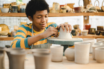 An Afro-haired potter sculpts a ceramic pot from clay on a potter's wheel. Art workshop concept, hands with clay making of a ceramic pot on the pottery wheel, hobby and leisure with pleasure concept - Powered by Adobe