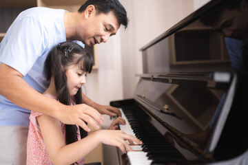 Fototapeta na wymiar Cute and focused young little Asian girl practicing songs and melody with father and tutor while pressing white and black key and creating new music on piano as hobby and dedication at home