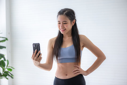 Portrait of smiling young Asian fit and healthy woman in sportswear taking selfie using smart phone camera against white background after workout and morning exercise ritual