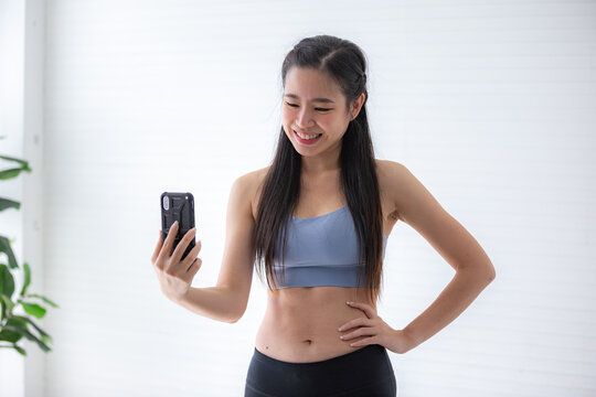 Portrait of smiling young Asian fit and healthy woman in sportswear taking selfie using smart phone camera against white background after workout and morning exercise ritual