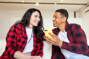 happy multiracial couple sitting on sofa at home and looking at smartphone, american guy and european girl using phone