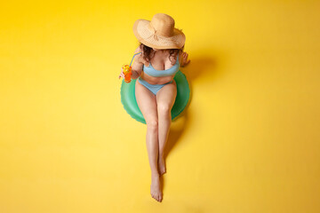 young girl in blue swimsuit sits on inflatable swimming ring holds orange alcoholic cocktail on yellow background