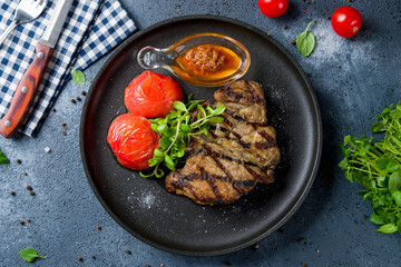 Fried pork loin with sauce and grilled tomatoes on black plate top view