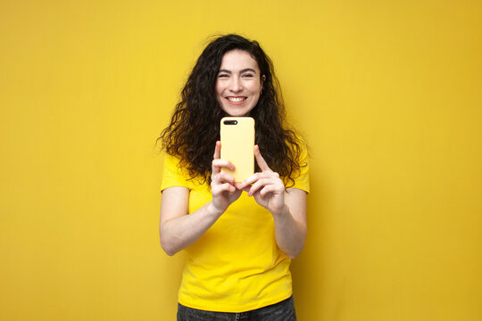 young cheerful curly girl in yellow t-shirt uses smartphone and smiles, brunette woman takes pictures on phone camera