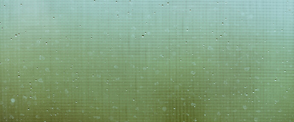 Mosquito net on wet window with rainy drops and dirt spots closeup. Rain droplets on glass with mosquito screen. Blurry minimal background with greenery bokeh against dirty window glass with raindrops