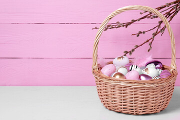 Wicker basket with festively decorated Easter eggs on white table against pink wooden background. Space for text
