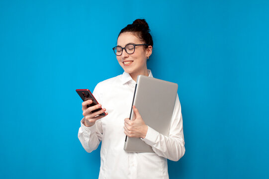 Successful Business Woman In White Shirt And Glasses Holds Laptop And Uses Smartphone On Blue Background