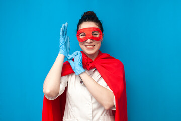 cheerful young girl doctor in uniform in superman costume on blue background, nurse superhero puts...