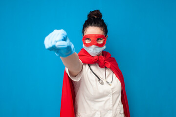 cheerful young girl doctor in uniform in superman costume on blue background, female nurse superhero shows forward