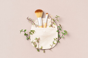 Eco-friendly cosmetic brushes in cotton bag and green leaves on beige background. Natural make up tools. Eco concept, creative.