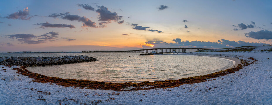 William T. Marler Bridge at Norriego Point in Destin Florida at sunset. Panoramic view of a sandy beach with Destin Bridge overlooking the scenic sky and orange horizon.