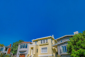 Homes with sunlit walls against blue sky viewed from the street in San Francisco. Facade of houses with balconies, outdoor stairs and bay windows.