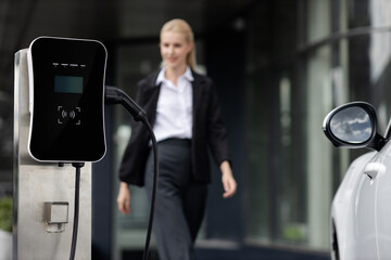 Focus public EV charging station for electric car with blurred progressive businesswoman walking in...