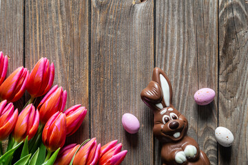 Easter background with a bouquet of pink tulips, a chocolate bunny and Easter eggs on a wooden background, top view, copy space.