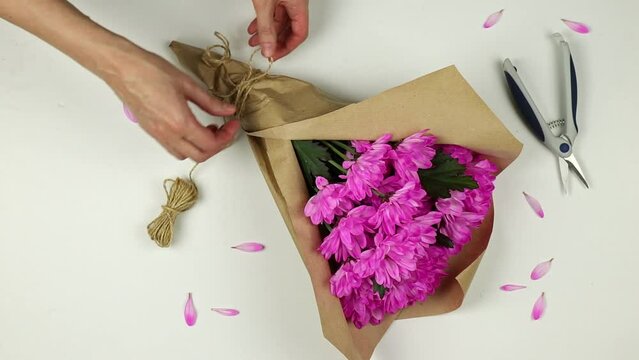 Women's hands cut jute rope with pruner on package of chrysanthemum bouquet on white desktop. Florist's table, flat lay.