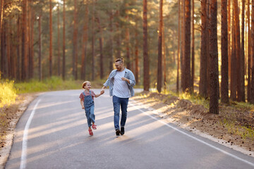 Dad and his little daughter have fun run along the forest road among tall pines. Family walk in the forest at sunset, a man and a little girl.