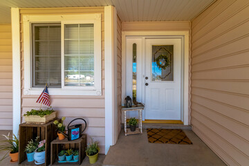 White front door with ornate glass panels near the ornamental flowers on pots on the side. There are decorative plants with small usa flag near the window on the left beside the door on the right.