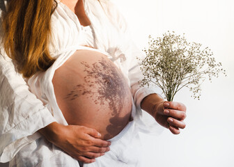 Cropped shot of unrecognizable pregnant woman holding beautiful flowers standing by white wall having shadows on belly.Lifestyle, minimalism concept.Pregnancy in spring,summer concept.