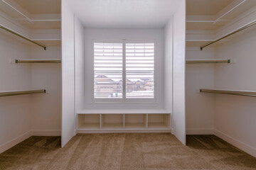 Carpeted walk-in closet with window blinds above the box shelves below. Walk-in closet interior with hanging rods on both sides of the window.