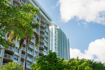 Multi-storey residential buildings with balconies and trees outdoors at Miami, Florida. There is a beige building on the left with steel railings near the building on the right with glass railings.