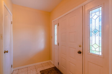 Front white door entrance interior with ornate glass on its sidelights. There is a white door on the left across the white front door on the right and a painted cream colored wall at the front.