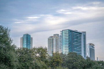 Fototapeta na wymiar Views of Austin, Texas cityscape from Butler Metro Park with trees at the front. Business and residential buildings with glass exterior against the cloudy sky background.