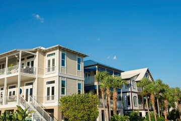 Three-storey houses with bushes and palm trees outdoors in Destin, Florida. There is a beige house at the front with stairs to the veranda under the balcony beside the house with blue trims.