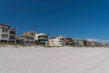 Fototapeta na wymiar White sand at the front of beach houses with footbridges over sand dunes in Destin, Florida. Row of houses with balconies and roof decks against the blue sky background.